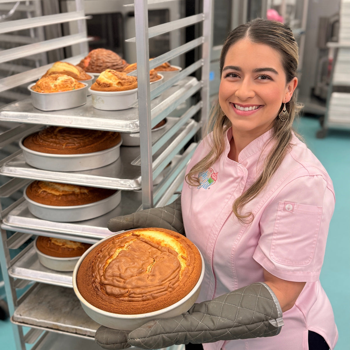 Chef Kristina Lavallee in a pink chef coat and gray oven mitts holds a freshly baked round cake in a white dish in a commercial kitchen, with metal baking racks filled with more cakes behind her.
